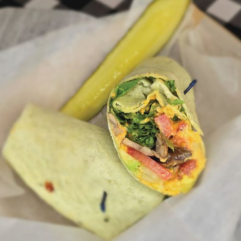 A deli wrap, cut in half, revealing colorful fillings, served with a pickle spear.